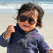 Scott is registered to the contest to win money with this photo: child, toddler, beach, ocean, waves, sand, sunglasses, portrait, outdoors, windy, hair, jacket, blue_clothing, face, expression, frown, hand, standing, sunlight, shoreline