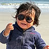 child, toddler, beach, ocean, waves, sand, sunglasses, portrait, outdoors, windy, hair, jacket, blue_clothing, face, expression, frown, hand, standing, sunlight, shoreline