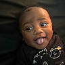 baby, child, tongue_out, black_clothing, cat_print, smiling, face, portrait, cute, infant, happy, expression, indoors, dark_background, closeup, person, head, young_child, playful, cozy