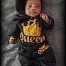 baby, child, infant, black_clothing, gold_crown, queen_text, hoodie, pants, bracelet, necklace, soft_surface, portrait, cute, young_child, fashion, laying_down, indoor, black_shoes, closeup, expression