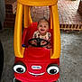 Raelynn is registered to the contest to win money with this photo: baby, child, toy_car, red, yellow, smiling, steering_wheel, plastic, playtime, outdoor, concrete, brick, happy, person, vehicle, wheels, garage, floor, cute, play