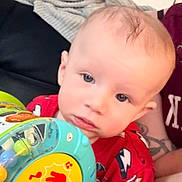 Noah joined the competition — help win amazing prizes! baby, child, toy, red_shirt, person, tattoo, hand, face, blue_eyes, indoor, couch, blanket, curious, music_toy, infant, skin, short_hair, holding, sitting, closeup