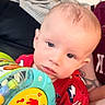 baby, child, toy, red_shirt, person, tattoo, hand, face, blue_eyes, indoor, couch, blanket, curious, music_toy, infant, skin, short_hair, holding, sitting, closeup