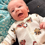Noah is registered to the contest to win money with this photo: baby, smiling, onesie, construction_vehicle_print, blanket, sofa, infant, happy, child, portrait, indoors, cute, young, fabric, clothing, person, face, lying_down, cozy, relaxed