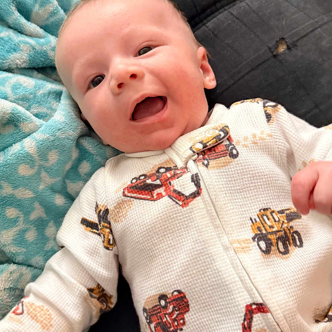 Noah is registered to the contest to win money with this photo: baby, smiling, onesie, construction_vehicle_print, blanket, sofa, infant, happy, child, portrait, indoors, cute, young, fabric, clothing, person, face, lying_down, cozy, relaxed