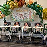 children, babies, infants, high_chairs, birthday_party, balloons, balloon_arch, jungle_theme, animal_decorations, elephant, tiger, giraffe, wall_art, indoor, celebration, colorful, sitting, portrait, festive, nursery