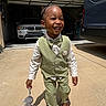 child, boy, smiling, vest, bow_tie, pants, toy, stuffed_animal, driveway, car, garage, sunlight, shadow, footwear, happy, outdoor, clothing, person, portrait, fashion