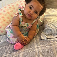 Kahlani is registered to the contest to win money with this photo: baby, child, curly_hair, dress, pink_bow, sock, bed, pillow, floral_pattern, cute, portrait, indoors, soft_lighting, cozy, toddler, smiling, hands, face, sitting, head_tilt
