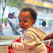 Melvin a rejoint le concours — aidez-le/la à gagner de superbes lots ! toddler, child, smiling, curly_hair, hoodie, white_clothing, red_chair, indoor, window, blurred_background, person, happy, young_child, face, playful, hands, seated, casual_clothing, daylight, candid