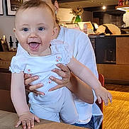 Artemis is registered to the contest to win money with this photo: baby, child, smiling, happy, person, indoor, table, wooden_table, holding, adult, face, blue_eyes, white_clothing, flower_pattern, hair, earrings, chair, floor, lamp, picture_frame