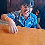 child, boy, glasses, smiling, blue_shirt, tropical_pattern, table, restaurant, booth, indoor, happy, person, seated, wood, furniture, casual, laughter, hair, face, clothing