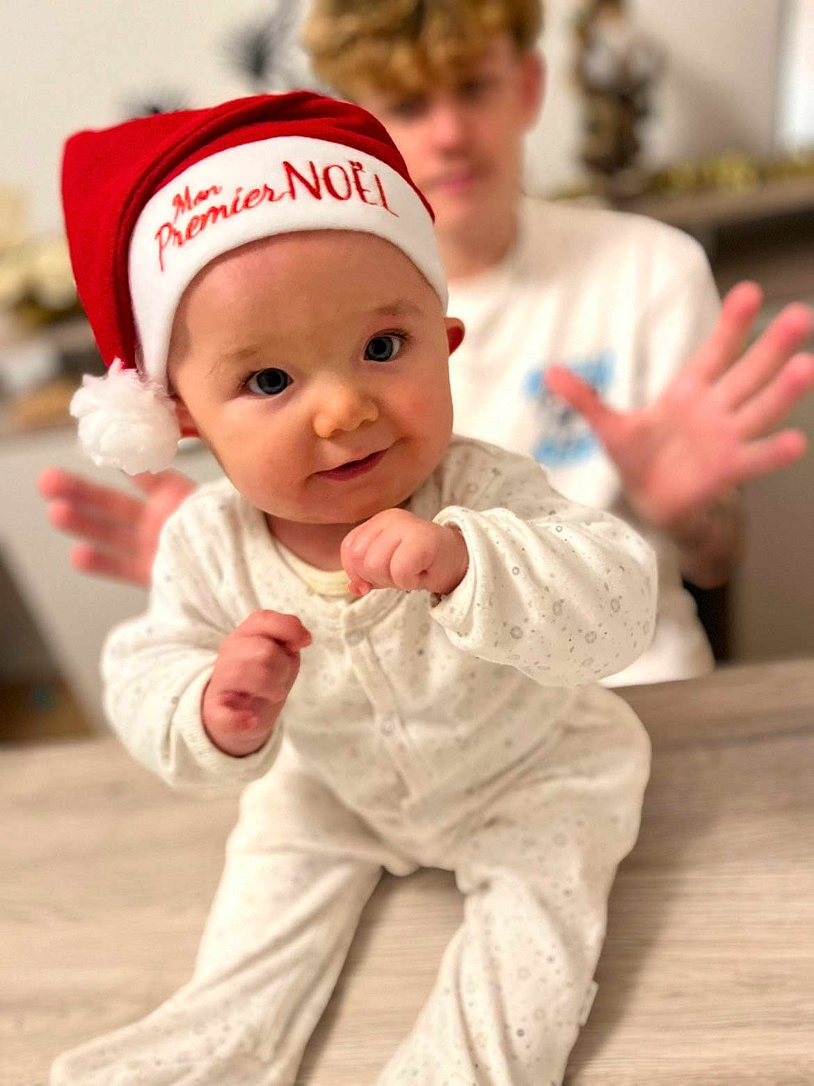 Elyo participe au concours pour gagner de l'argent avec cette photo : baby, child, santa_hat, holiday, christmas, indoor, table, white_clothing, portrait, cute, smile, festive, person, face, blurry_background, hands, wooden_surface, happy, infant, family