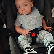Mason is registered to the contest to win money with this photo: baby, child, car_seat, bib, striped_pants, sneakers, indoor, seated, curious_expression, clothing, footwear, person, young_child, cute, infant, safety_buckle, head, face, hands, legs