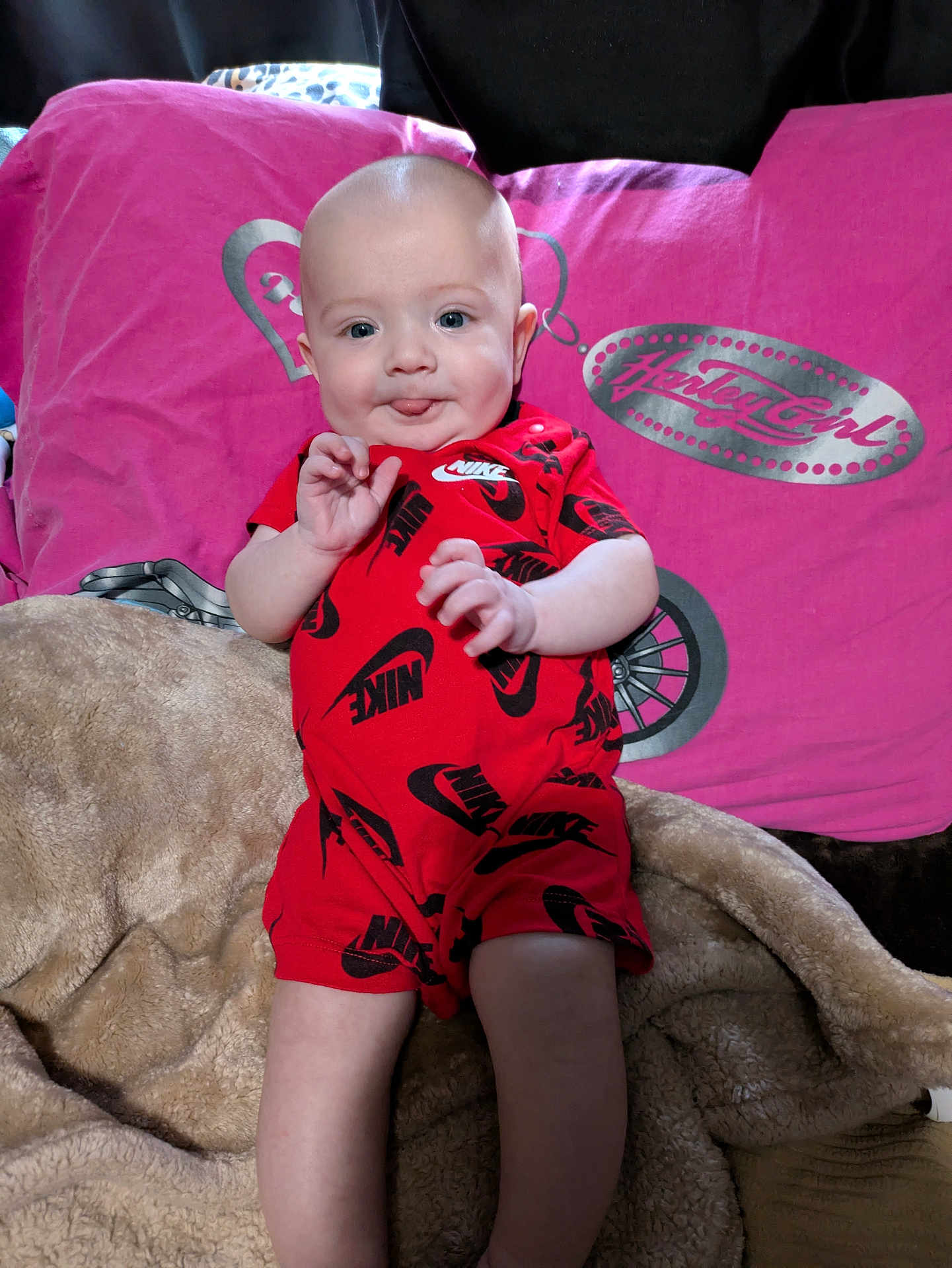 Anakin is registered to the contest to win money with this photo: baby, infant, red_clothing, nike, blanket, pillow, pink, beige, face, tongue_out, smile, sitting, indoor, cute, child, person, soft_texture, cozy, happy, playful