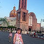 Mia is registered to the contest to win money with this photo: child, girl, rock, church, tower, building, street, sky, daytime, dress, sandals, backpack, people, urban, sidewalk, tree, architecture, outdoor, walking, public