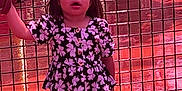 Mia is registered to the contest to win money with this photo: child, girl, holding_hand, floral_top, purple_pants, sneakers, fence, amusement_park, playground, metal_fence, outdoor, daylight, standing, looking_up, casual_clothing, person, photography, colorful, curly_hair, bow