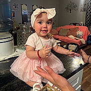 Betzaida Nairobi joined the competition — help win amazing prizes! baby, bow, child, cushions, cute, glass_jar, hand, happy, headband, home, indoor, kitchen_counter, living_room, person, pink_dress, playful, smiling, sofa, toddler, water_dispenser