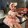 baby, bow, child, cushions, cute, glass_jar, hand, happy, headband, home, indoor, kitchen_counter, living_room, person, pink_dress, playful, smiling, sofa, toddler, water_dispenser