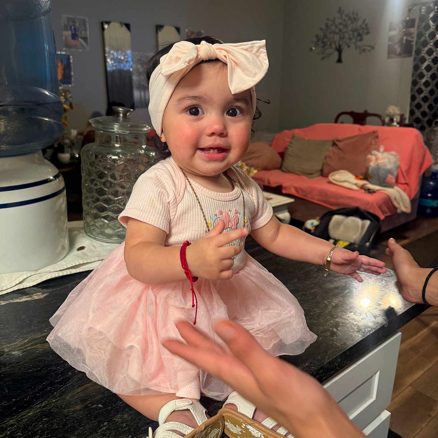 Betzaida Nairobi joined the competition — help win amazing prizes! baby, bow, child, cushions, cute, glass_jar, hand, happy, headband, home, indoor, kitchen_counter, living_room, person, pink_dress, playful, smiling, sofa, toddler, water_dispenser