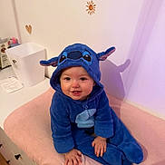 Romy a rejoint le concours — aidez-le/la à gagner de superbes lots ! baby, child, blue_onesie, stitch_costume, smiling, cute, indoor, pink_surface, decorated_wall, heart_decal, sun_decal, happy, person, infant, soft_texture, seated, hand, face, hooded_clothing, playful