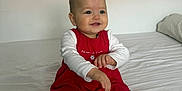 Romy a rejoint le concours — aidez-le/la à gagner de superbes lots ! baby, child, smiling, red_dress, white_shirt, white_tights, heart_pattern, bed, pillow, indoor, cute, sitting, happy, young_child, portrait, infant, person, human, cozy, casual
