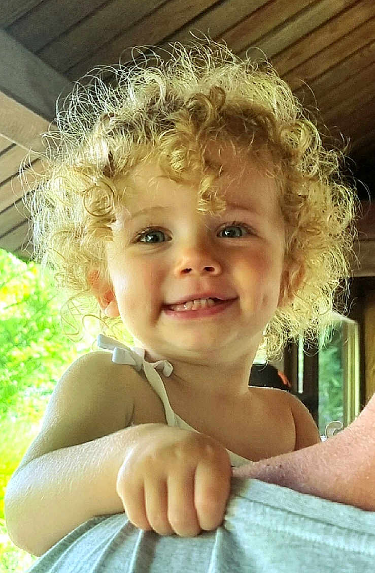 Angel Agathe a rejoint le concours — aidez-le/la à gagner de superbes lots ! toddler, child, curly_hair, smile, dimples, face, portrait, indoor, wooden_ceiling, greenery, person, happy, holding, baby_clothes, skin, hand, cute, bright, natural_light, close_up