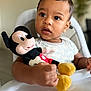 toddler, child, plush_toy, mickey_mouse, high_chair, indoor, curious, expression, clothing, person, face, hand, toy, seat, baby, closeup, portrait, soft_focus, furniture, cute