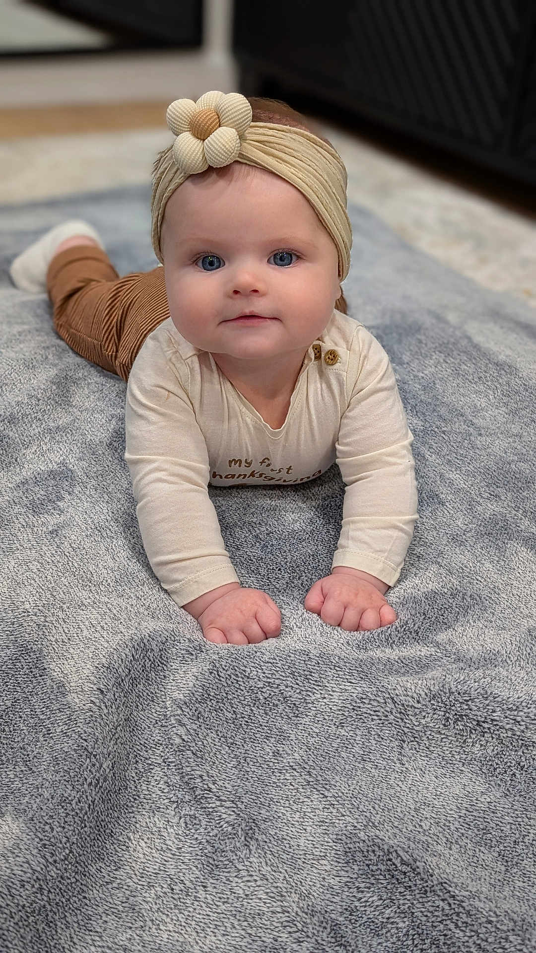 Victoria is registered to the contest to win money with this photo: baby, child, headband, flower, blue_eyes, blanket, soft_texture, indoor, infant, cute, curious, long_sleeve, brown_pants, portrait, hands, floor, cozy, toddler, lying_down, close_up