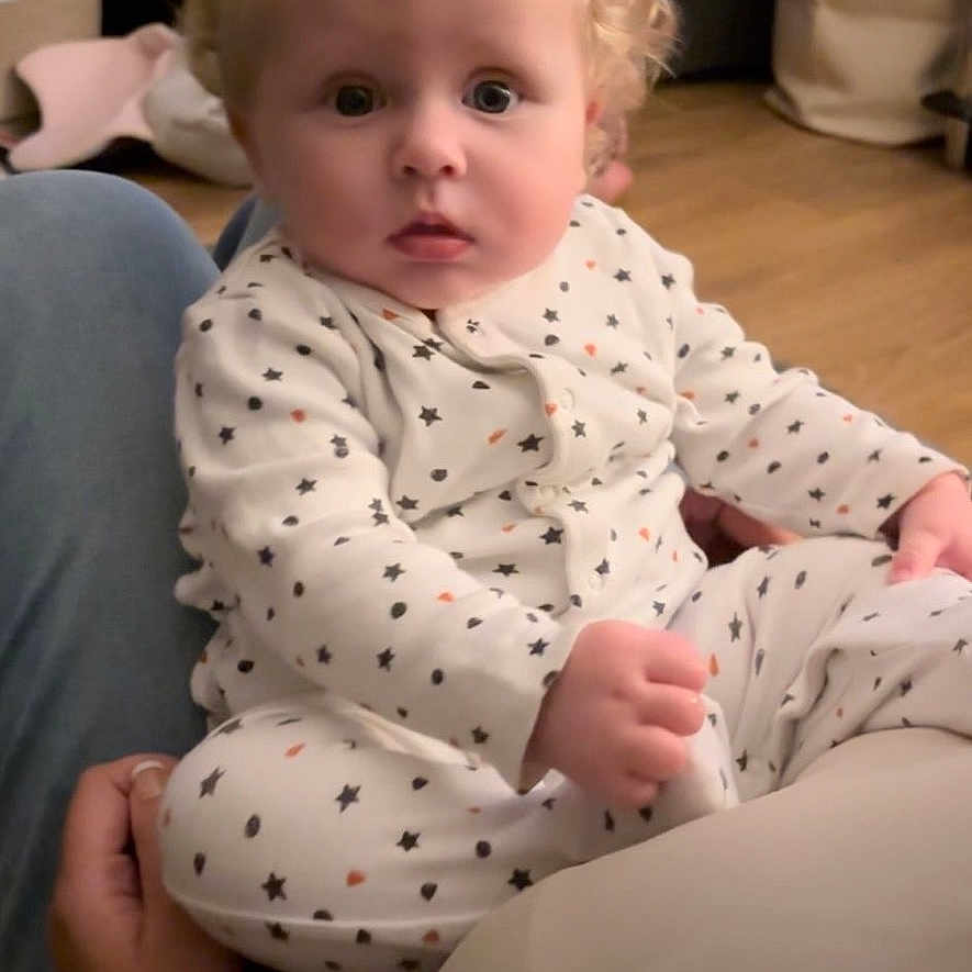 Noah joined the competition — help win amazing prizes! baby, blonde_hair, child, clothing, cozy, curly_hair, cute, expression, eyes, face, floor, hand, indoor, infant, lap, onesie, person, sitting, stars_pattern, wooden_floor