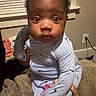 baby, infant, pajamas, blanket, indoor, face, person, child, pacifier, curly_hair, expression, cute, seated, window_blinds, plug_socket, soft_toy, home, sleepwear, carpet, curious