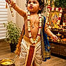 child, toddler, portrait, traditional_clothing, indian_culture, religious_decoration, peacock_feather, flute, garland, jewelry, curly_hair, makeup, barefoot, smiling, indoor, altar, oil_lamps, flowers, butter, festive