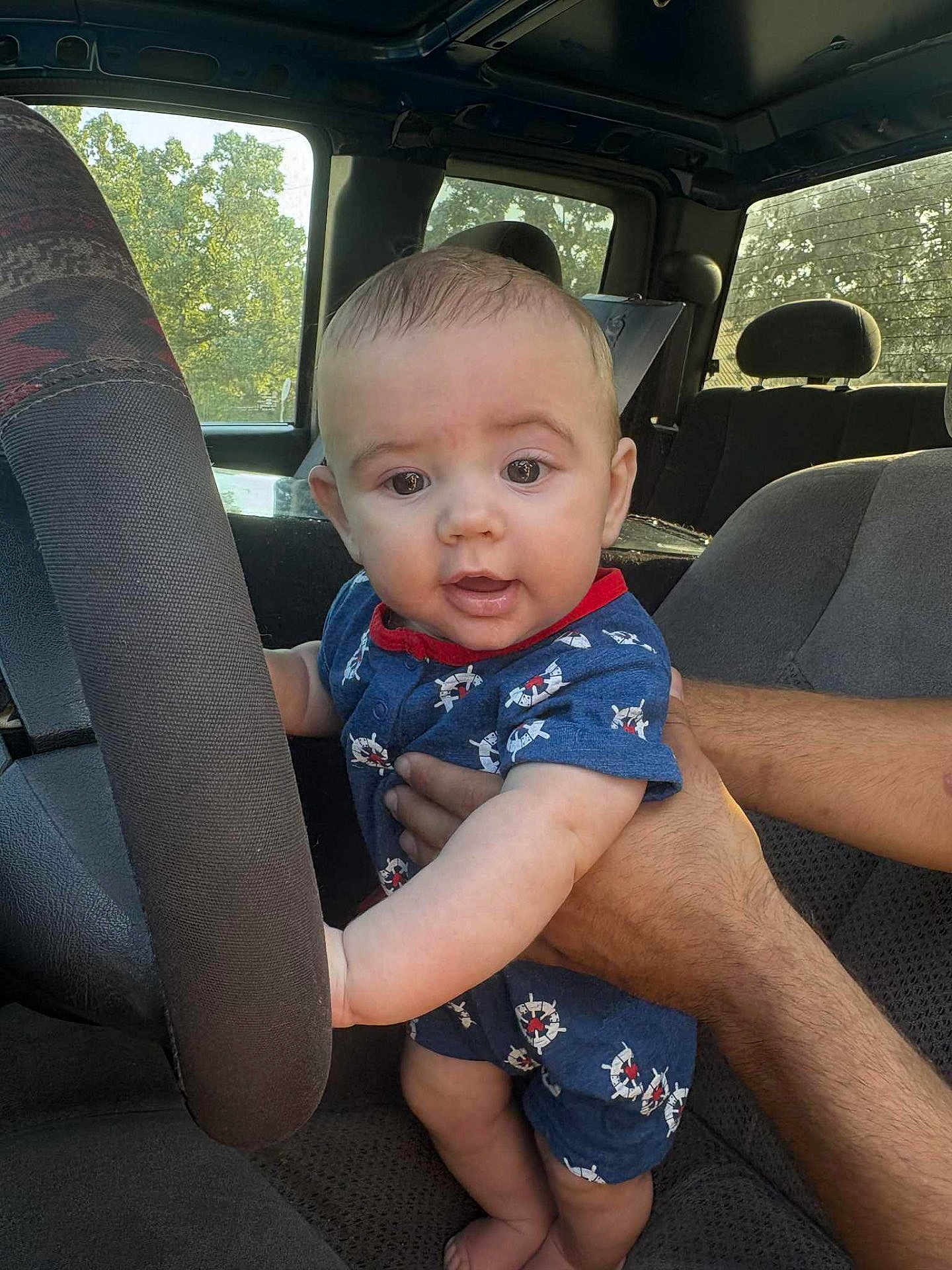 Kingston is registered to the contest to win money with this photo: baby, child, infant, car_interior, steering_wheel, hand, blue_clothing, nautical_pattern, seat, window, sunlight, person, cute, curious, holding, arm, head, eyes, expression, vehicle