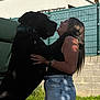 dog, woman, outdoor, grass, fence, building, smile, hug, black_dog, jeans, tank_top, sunlight, collar, long_hair, watch, affection, shadow, greenery, happy, casual