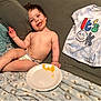 toddler, child, smiling, couch, blanket, plate, food, messy_face, socks, shirt, indoor, cozy, happy, relaxed, baby, person, clothing, furniture, home, casual