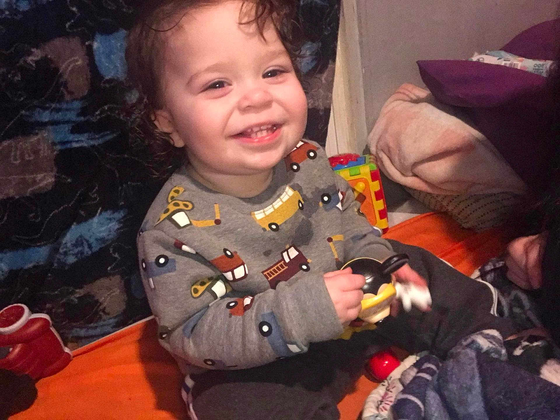 Brayden joined the competition — help win amazing prizes! child, toddler, smile, curly_hair, toy, sitting, blanket, pillow, indoor, clothing, sweatshirt, pants, happy, face, person, play, colorful, comfort, home, cozy