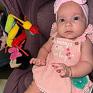 Valerie is registered to the contest to win money with this photo: baby, child, car_seat, pink_dress, headband, pacifier, plush_toys, bracelet, indoor, cute, infant, person, young_child, seat, accessory, soft_toys, closeup, portrait, expression, colorful