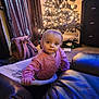 Octavia is registered to the contest to win money with this photo: child, baby, toddler, christmas_tree, christmas_lights, ornament, living_room, pink_sweater, backpack, curtain, couch, leather_sofa, carpet, holiday, indoor, cozy, portrait, sitting, curious_expression, floor_mat
