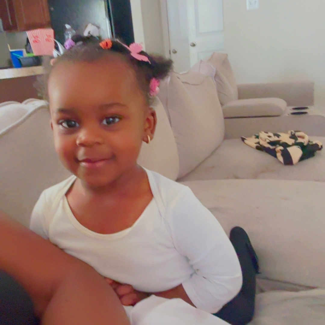 Kaylee is registered to the contest to win money with this photo: child, toddler, smile, hair_clips, living_room, couch, sofa, home_interior, person, indoors, furniture, kitchen, cabinet, framed_picture, door, white_shirt, cute, happy, portrait, casual