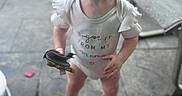 Bria is registered to the contest to win money with this photo: toddler, child, barefoot, onesie, toy, outdoor, patio, curious, standing, wide_eyes, ruffled_sleeves, blurry_background, hair, expression, footwear_none, concrete_floor, furniture, sunlight, person, young
