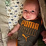 Bryson is registered to the contest to win money with this photo: baby, infant, pacifier, blanket, patterned_blanket, clothing, onesie, puma, logo, face, head, skin, arm, leg, person, indoor, cute, expression, lying_down, baby_garment