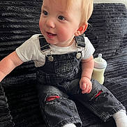 Keon is registered to the contest to win money with this photo: toddler, baby, child, overalls, white_tshirt, couch, black_couch, baby_bottle, sitting, indoors, light_hair, ripped_jeans, smiling, cute, portrait, face, happy, cozy, furniture, casual_clothing