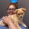 person, dog, puppy, glasses, smile, hugging, indoor, blue_shirt, curly_hair, wall_art, painting, animal, pet, cute, fur, hand, face, happy, portrait, closeup