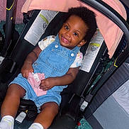 Kaylani is registered to the contest to win money with this photo: toddler, child, car_seat, denim_overalls, socks, indoor, smiling, baby, happy, seatbelt, pink_cloth, floor, furniture, head_support, curly_hair, playpen, cushion, clothing, person, portrait