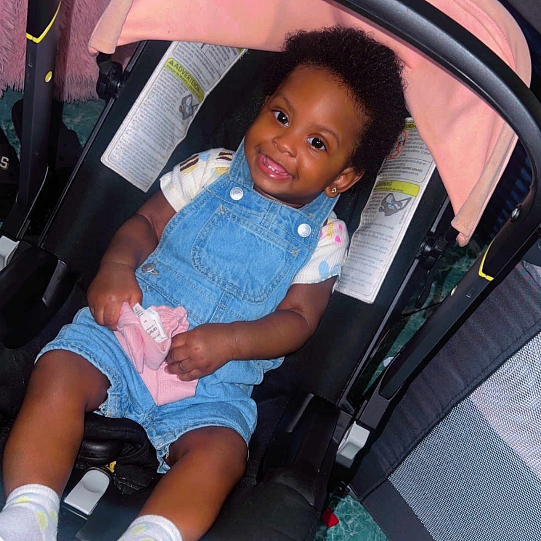 Kaylani is registered to the contest to win money with this photo: toddler, child, car_seat, denim_overalls, socks, indoor, smiling, baby, happy, seatbelt, pink_cloth, floor, furniture, head_support, curly_hair, playpen, cushion, clothing, person, portrait