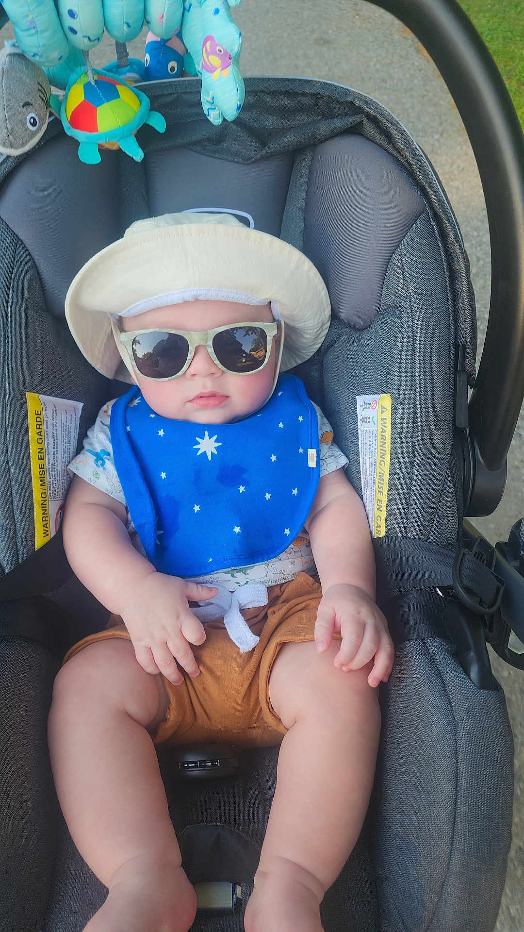 Frederick joined the competition — help win amazing prizes! baby, stroller, sunglasses, hat, bib, blue, brown_shorts, toy, hanging_toys, outdoor, child, infant, cute, summer, sun_protection, seatbelt, car_seat, person, legs, hand