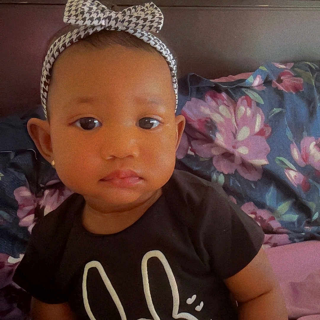 Tyanna is registered to the contest to win money with this photo: baby, bed, black_shirt, bow, bunny_graphic, child, clothing, cute, earrings, floral_bedspread, headband, indoor, person, pillow, portrait, serious_expression, sitting, toddler, wooden_headboard, young_child