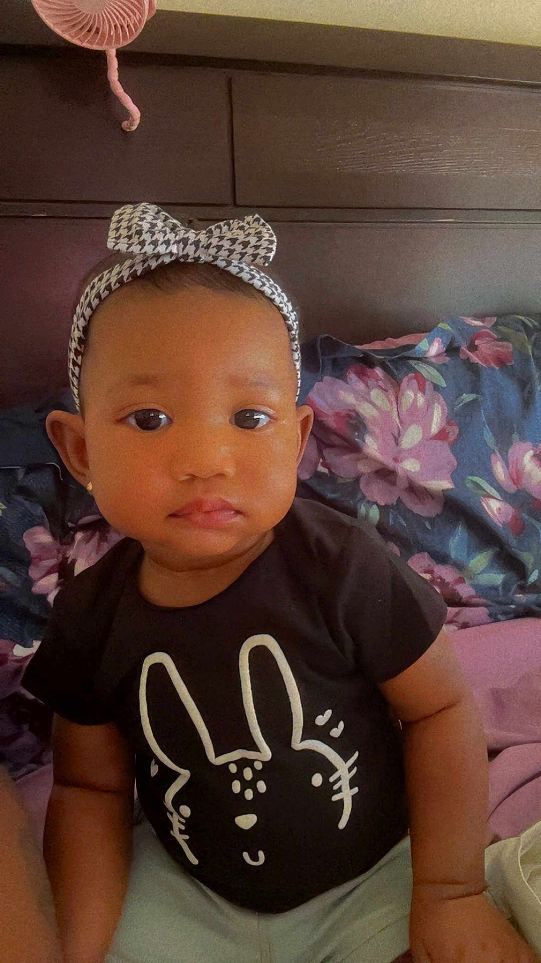 Tyanna is registered to the contest to win money with this photo: toddler, child, baby, headband, bow, black_shirt, bunny_graphic, floral_bedspread, bed, pillow, wooden_headboard, indoor, portrait, cute, serious_expression, person, clothing, sitting, young_child, earrings