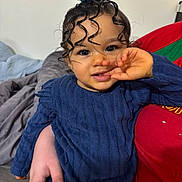 Maëna a rejoint le concours — aidez-le/la à gagner de superbes lots ! child, toddler, baby, portrait, curly_hair, top_knot, blue_outfit, hand_near_mouth, smile, big_eyes, indoor, blanket, pillow, bed, person, adult_hand, red_shirt, long_sleeves, skin, closeup