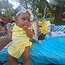 baby, child, yellow_outfit, headband, outdoor, picnic, table, blue_tablecloth, soda_can, coloring_sheet, tree, grass, hand, person, sunlight, daylight, food, drink, family, summer