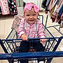 Lola is registered to the contest to win money with this photo: baby, child, shopping_cart, pink_bow, headband, plaid_shirt, dark_pants, white_socks, indoor, store, clothing_rack, blanket, happy, smiling, holding, sitting, retail, cute, floor, person